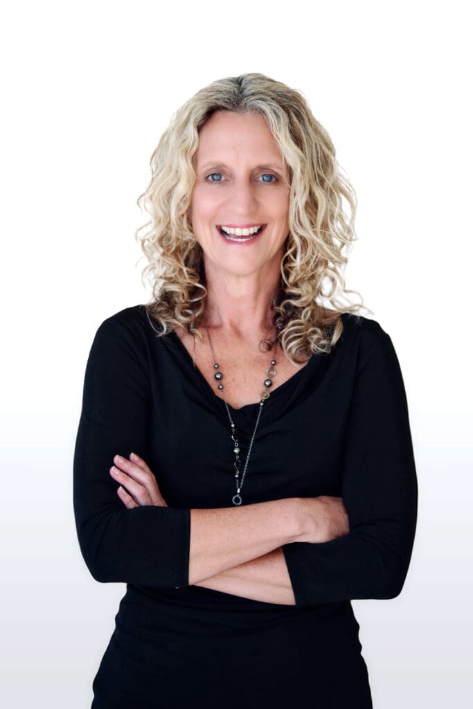 A woman with curly blonde hair, wearing a black long-sleeve top and a necklace, stands smiling with her arms crossed against a plain white background.