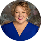 A smiling woman with short curly blonde hair, wearing a royal blue V-neck top and statement earrings, poses in front of a softly blurred floral background.