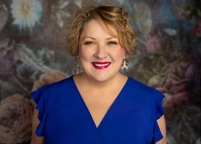 A woman with short, curly blonde hair wearing a bright blue dress and silver earrings smiles in front of a floral, blurred background.